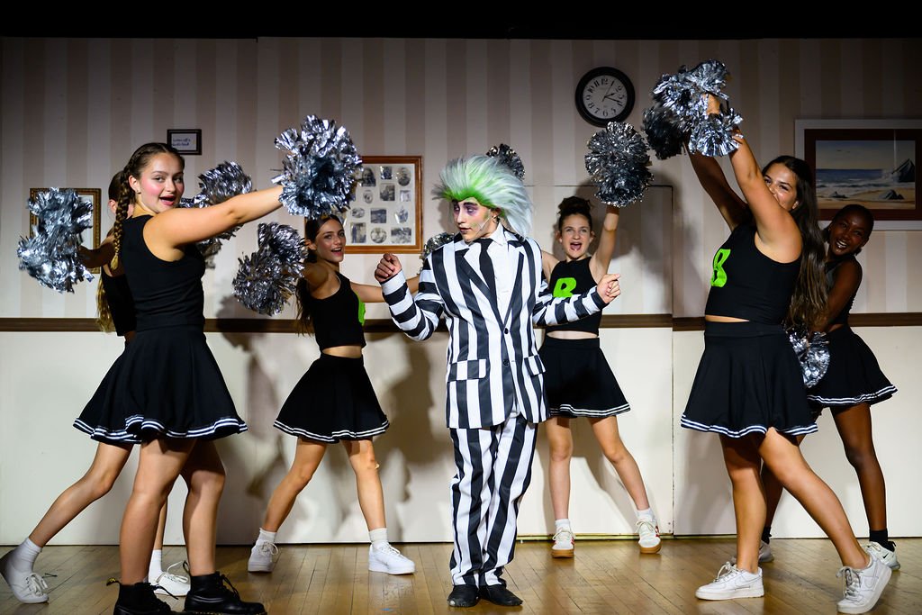 A group of five young girls and a clown performer in a costume with black and white stripes, holding pom-poms, dancing in a room with a striped wallpaper, framed pictures, and a clock on the wall.