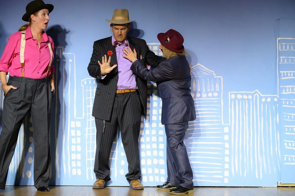 Three people on stage engaged in a theatrical performance. The woman on the left wears a pink shirt, gray pants, suspenders, and a black hat, looking surprised. The man in the center wears a pinstripe suit, a purple shirt, a bow tie with pink and pur
