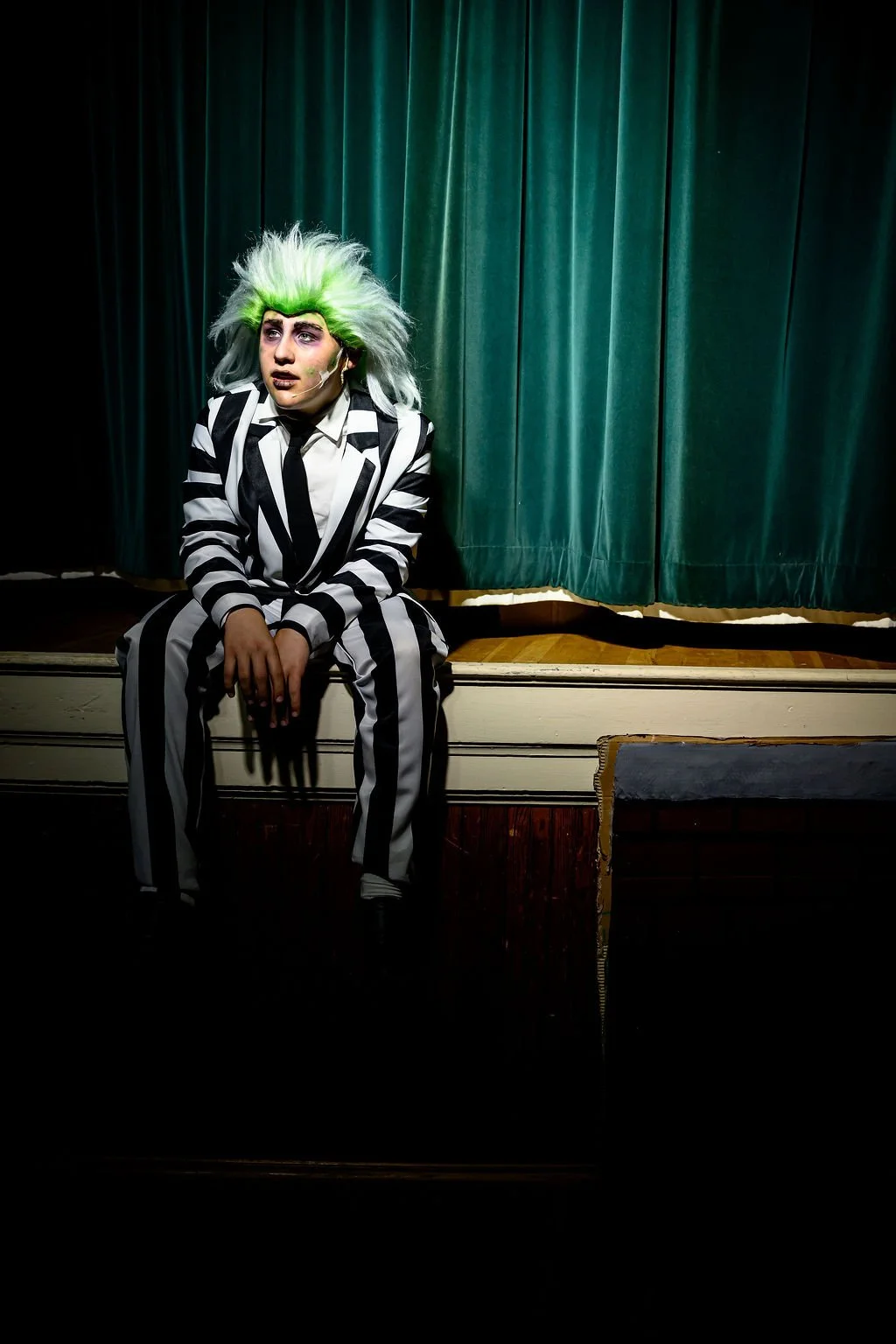 A person dressed as Beetlejuice in a black and white striped suit with wild green and white hair, sitting on the edge of a stage with green curtains in the background.