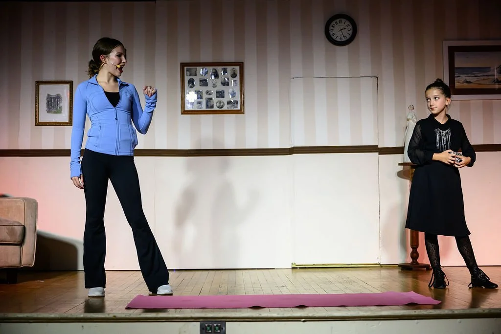 Two young girls on stage, one in a blue zip-up jacket and black pants, speaking or performing, and the other in a black dress with decorative details, holding a camera, in a room with framed pictures and a clock on the wall.