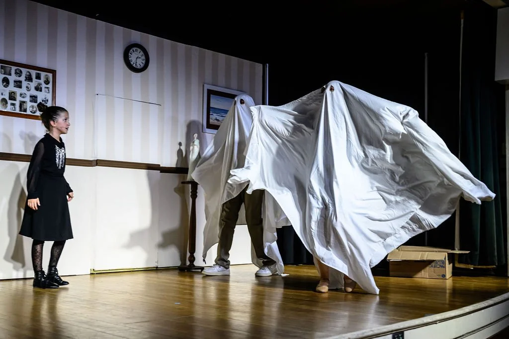 Children performing a play on stage with a person hiding behind a white sheet acting as a ghost