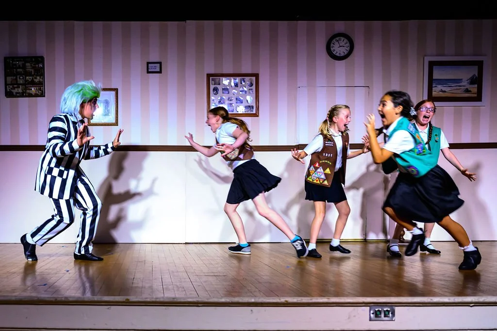 Children performing in a school play on a stage with striped wallpaper and framed pictures.