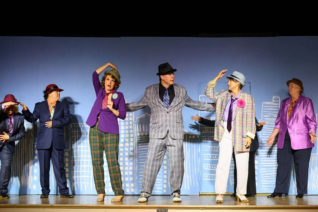 Group of actors performing on stage, dressed in colorful vintage clothing, with a cityscape painted backdrop.