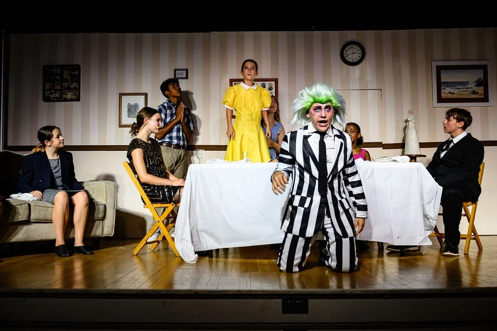 Children performing in a play, with one dressed as Beetlejuice in a black-and-white striped suit and green hair, others seated or standing around a table and sofa in a room with striped wallpaper and framed pictures.