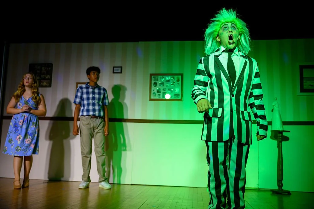 A theatrical scene with two young actors, a girl in a blue floral dress and a boy in a checkered shirt, standing on stage with a person wearing a green and black striped suit and wig, in a room decorated with framed pictures and green lighting.