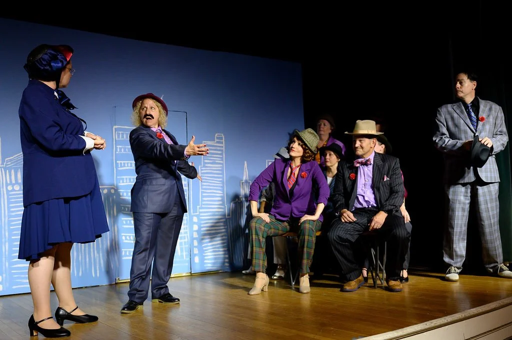 A theatrical play with actors in costumes on stage, featuring a woman in a blue suit and hat speaking to a woman in a purple blazer, with others seated and standing nearby.