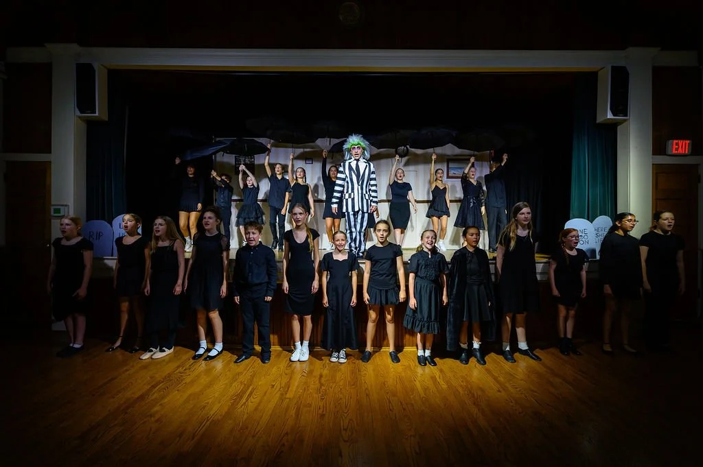 A group of children performing on stage, with some dressed as characters from Beetlejuice in a theatrical production.