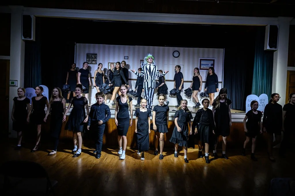 Children performing a school play on stage with a clown, some children are standing in a semi-circle in front, and others are dancing or standing in the background, with signs reading 'RIP' and 'Gone Here She Lies' in the background.
