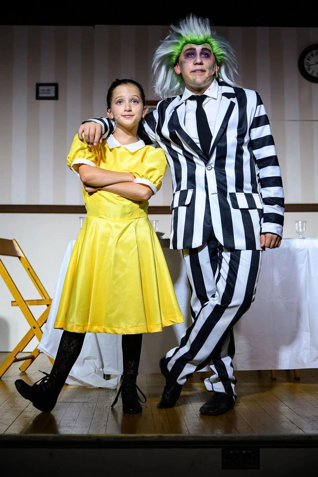A young girl in a yellow dress and a man dressed as Beetlejuice, in a black-and-white striped suit and wild green-topped hair, stand together on stage with their arms around each other.