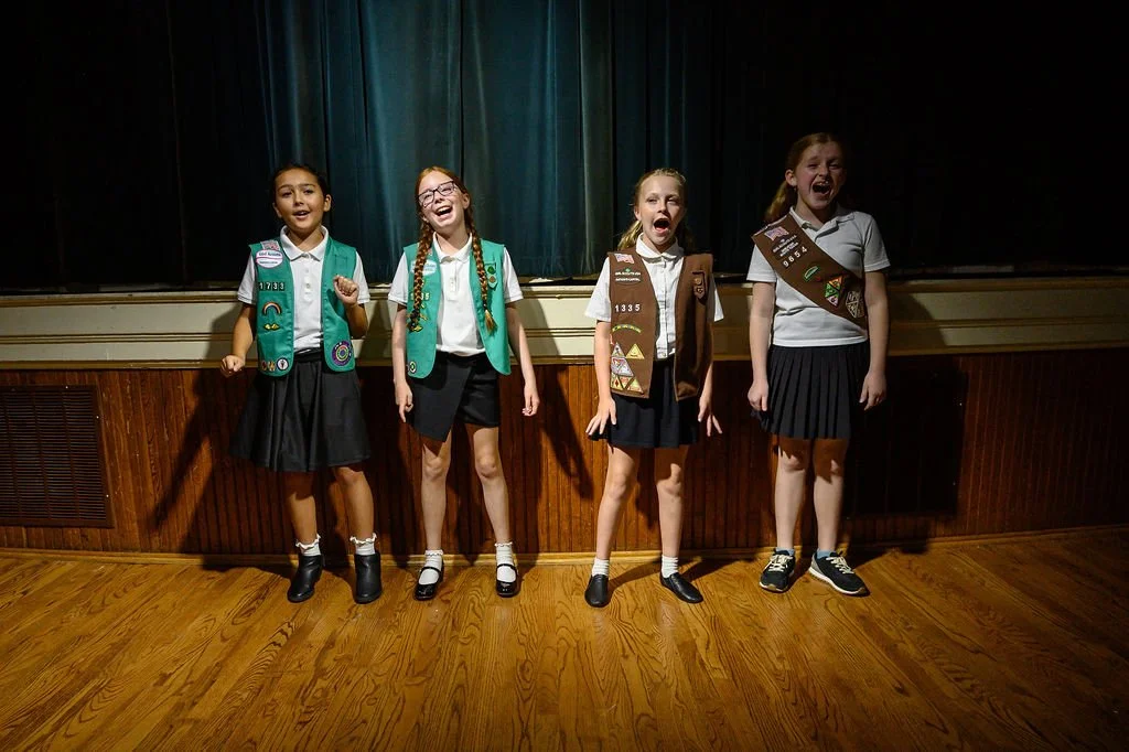 Four girl scouts in uniform standing on a stage, smiling and singing.