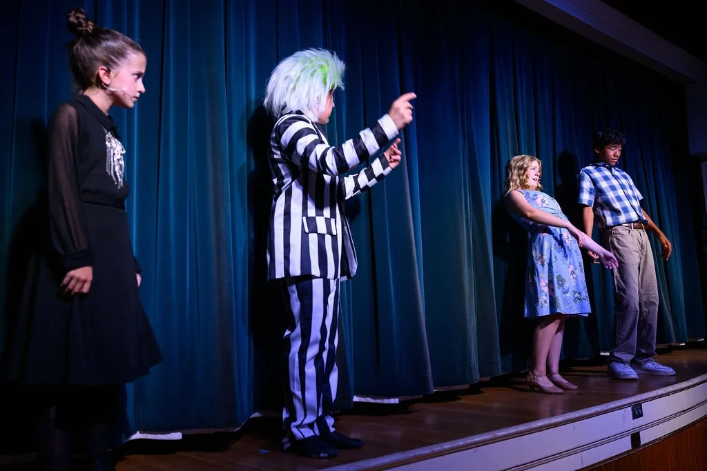 Children performing on stage with a blue curtain backdrop, dressed in costumes, including a girl in a black dress, a person in a striped outfit with green hair, a girl in a floral dress, and a boy in plaid shirt and khakis.