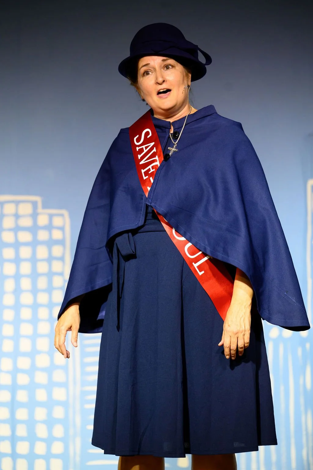 Woman on stage wearing a blue outfit, a black hat, and a red sash