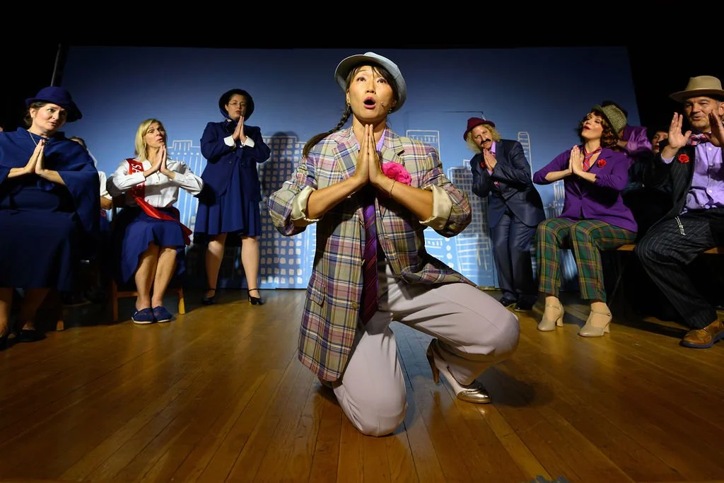 A theatrical performance with actors dressed in colorful costumes, one actor kneeling center stage with hands pressed together in a prayer-like gesture, surrounded by other actors seated and standing against a cityscape background.