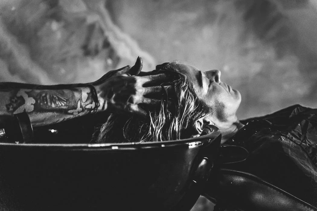 A person with long hair in a sink being washed or rinsed off by another person with tattooed arm, in black and white.