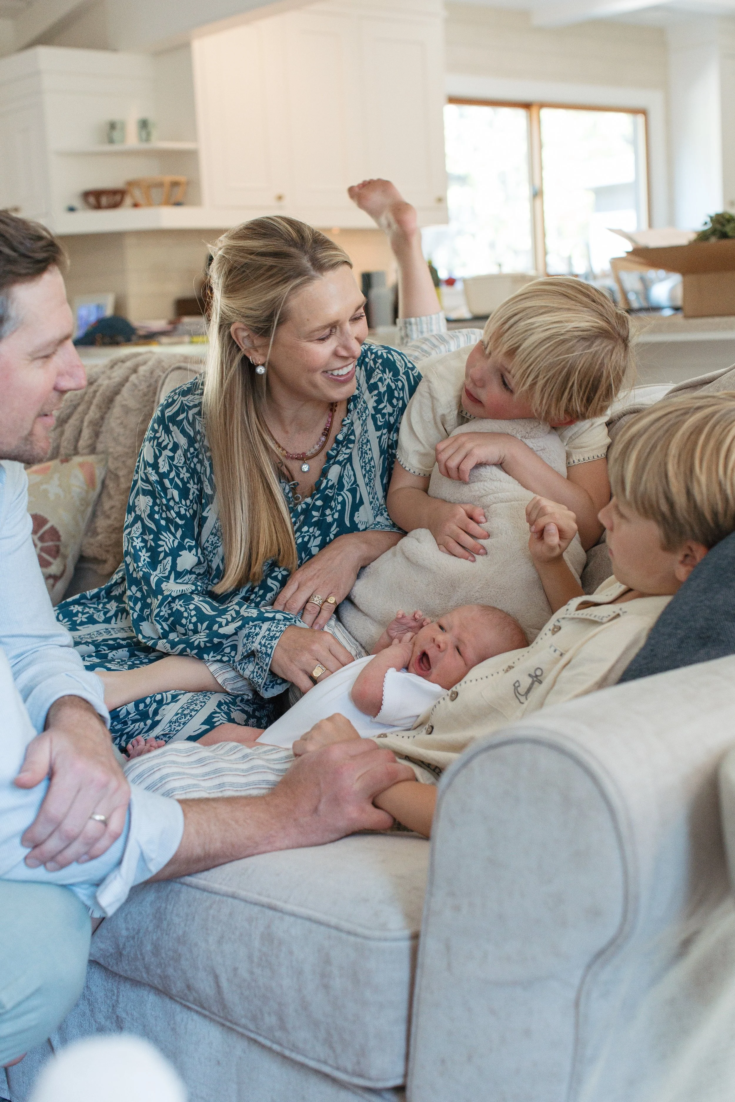 A family gathered on a beige couch in a living room, celebrating the birth of a newborn. The mother, with long blonde hair, is smiling warmly at the baby. The father is sitting nearby, holding the newborn, while other children and family members look