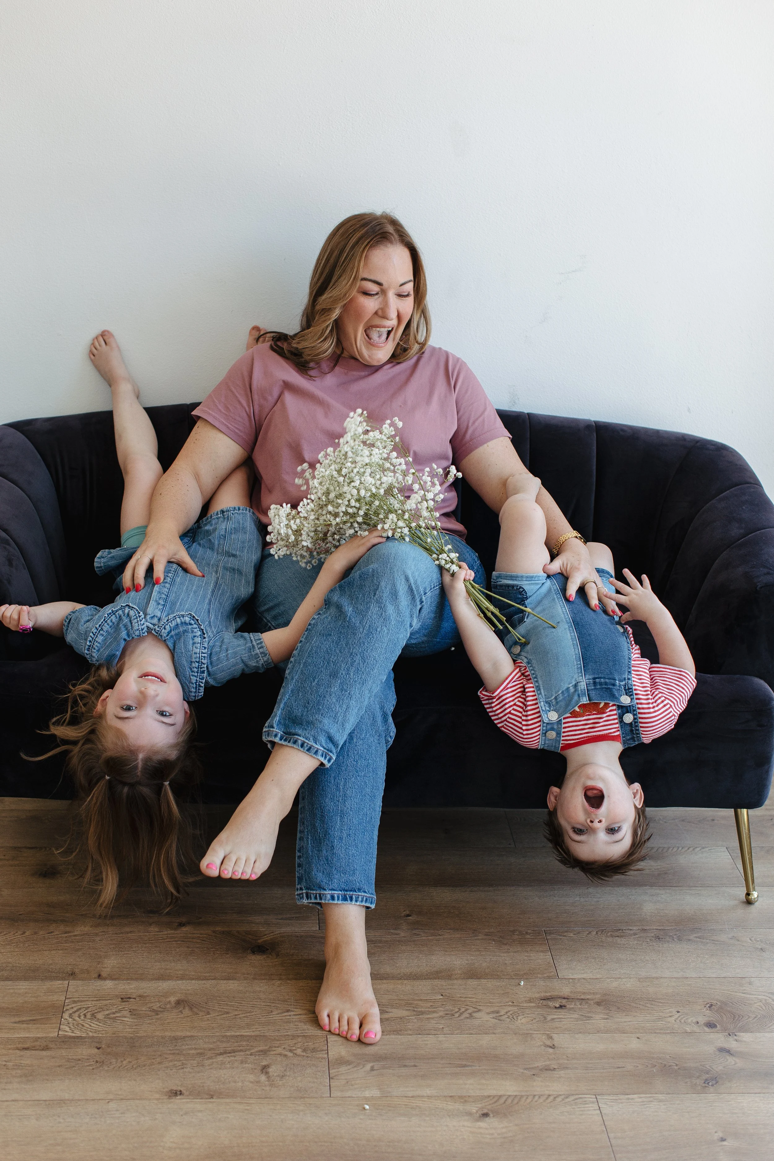 A woman sitting on a black sofa with four children, holding a bouquet of white flowers. Two children are upside down on the sofa, one with long hair hanging down, faces showing joy and playfulness.