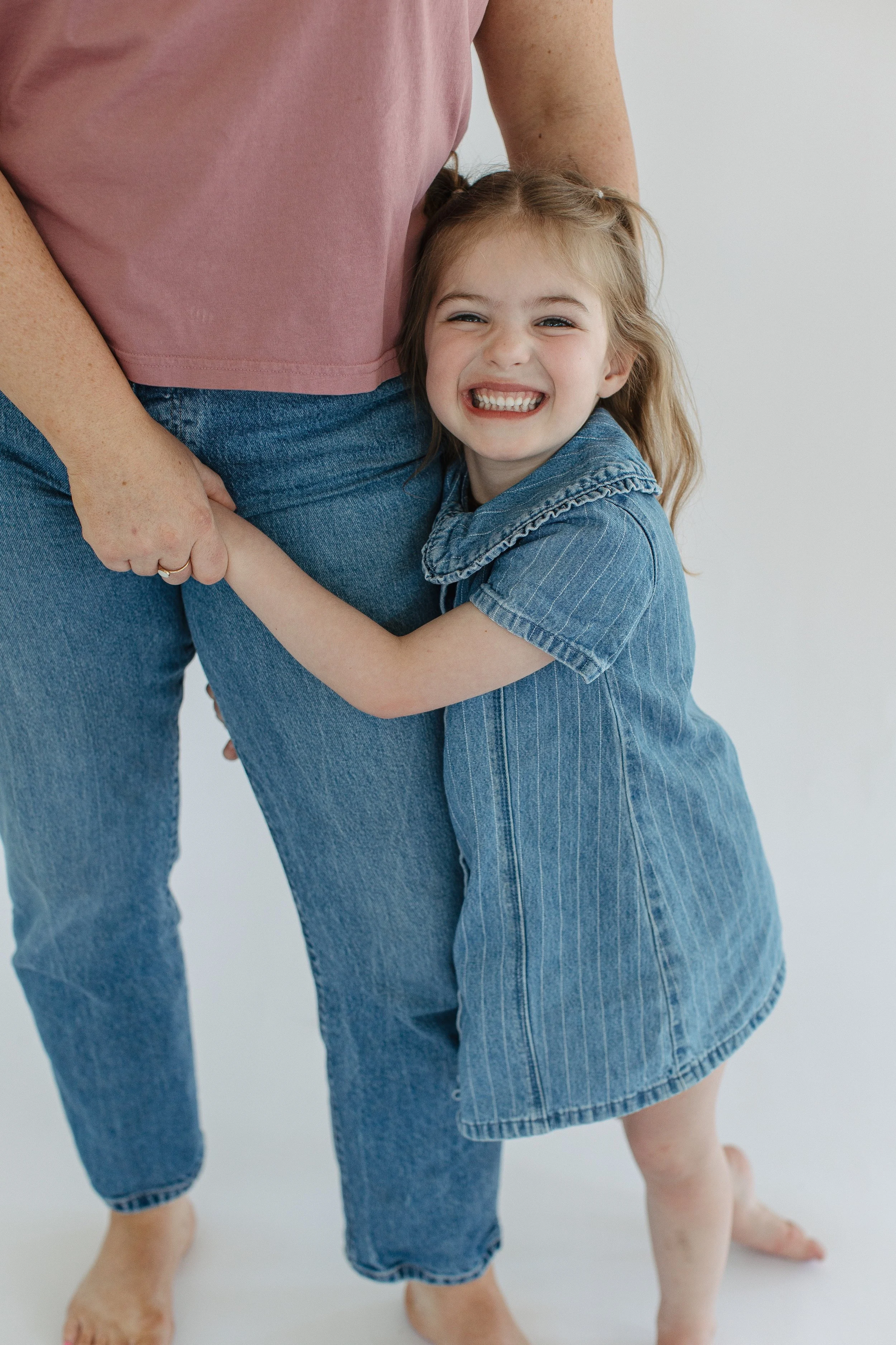A young girl hugging an adult, both wearing blue jeans. The girl is smiling widely, showing her teeth, and wearing a blue denim dress. The adult's upper body and legs are visible, with one hand resting on the girl's arm.