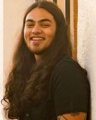 Young man with long, dark hair smiling and wearing a black shirt.