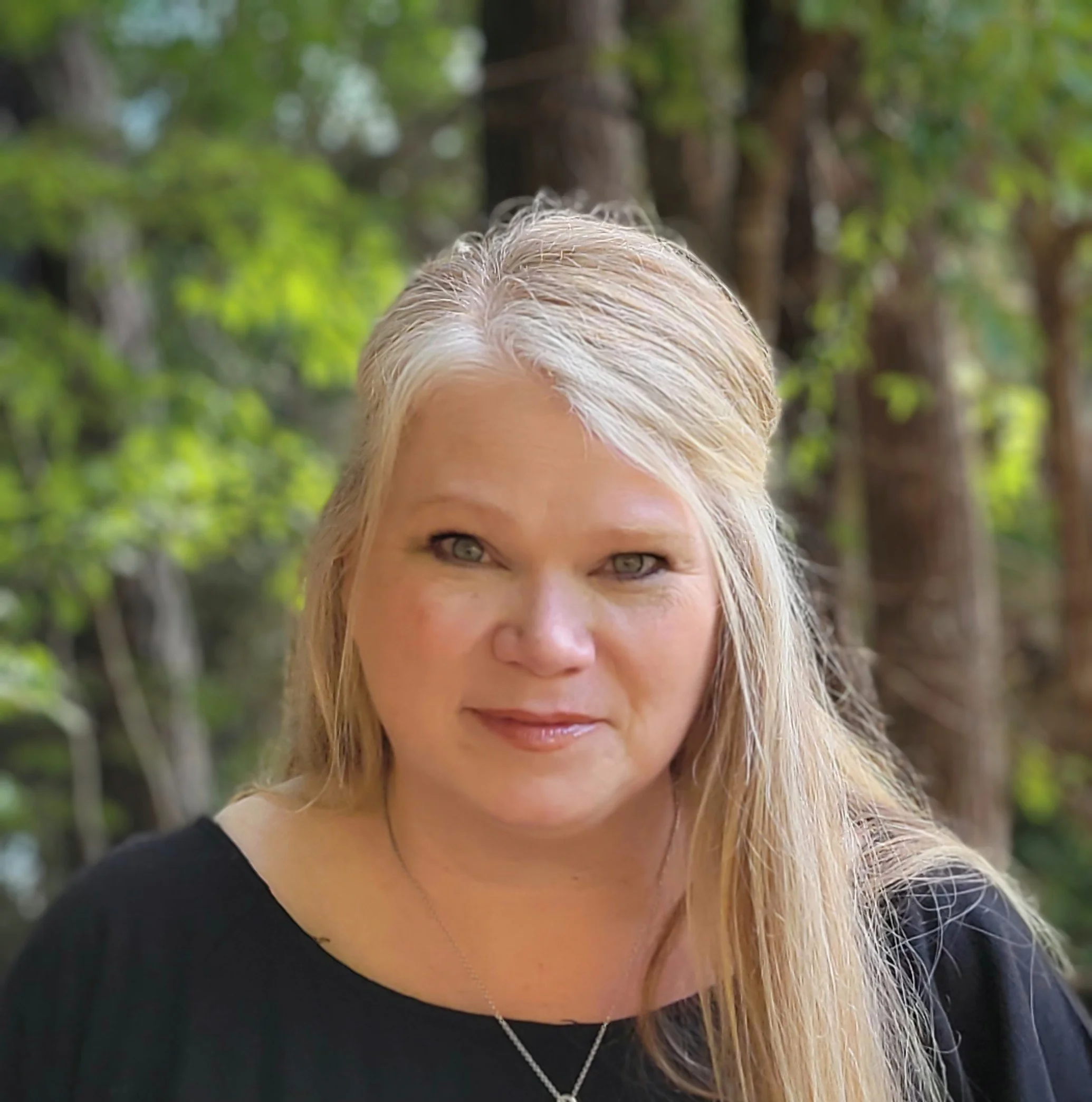 Portrait of a woman with blonde hair and fair skin, wearing a black top, outdoors with a forest background.