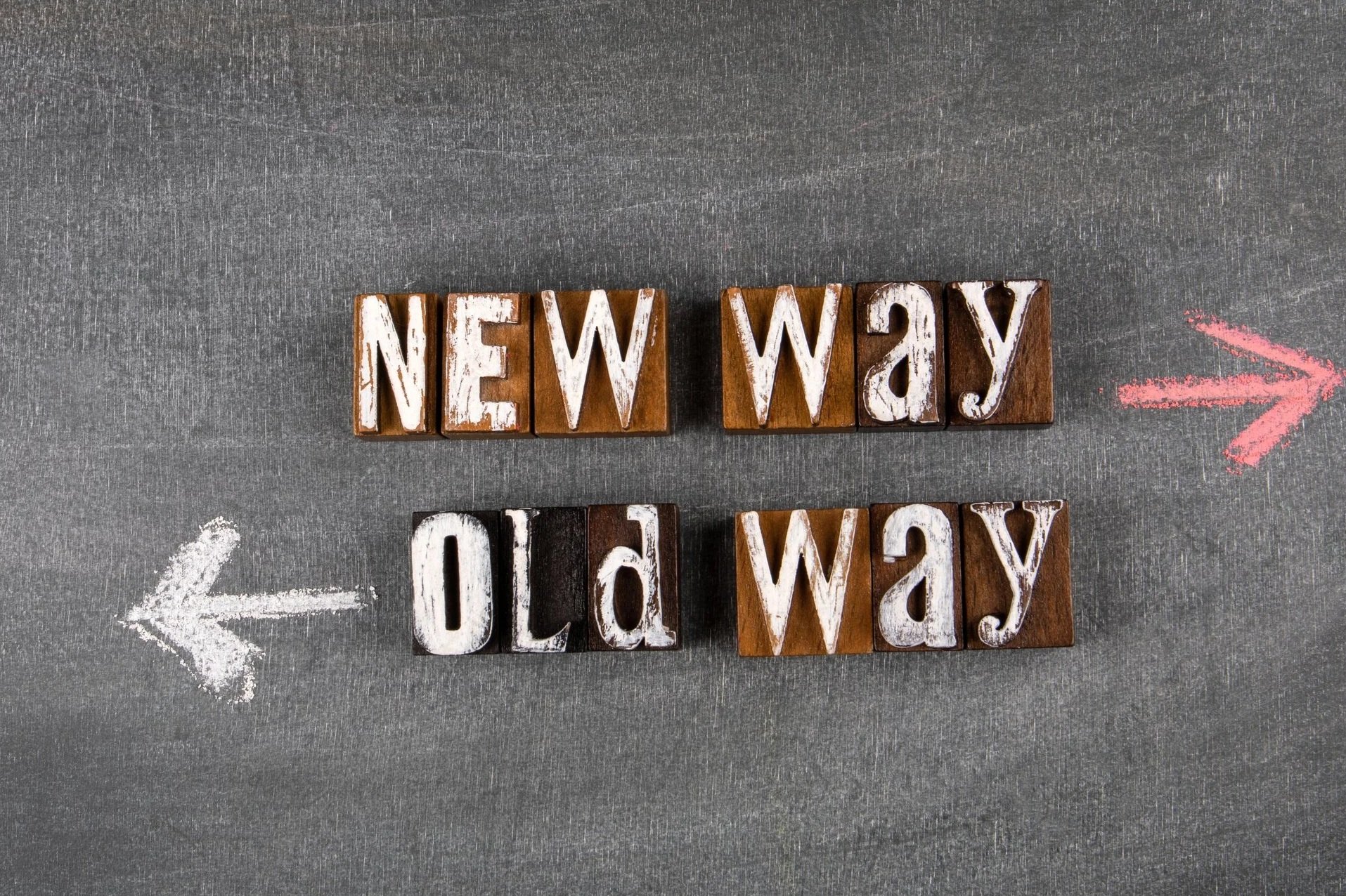 Wooden letter blocks displaying the words 'New Way' pointing to the right and 'Old Way' pointing to the left, with pink and white chalk arrows indicating directions on a gray textured surface.