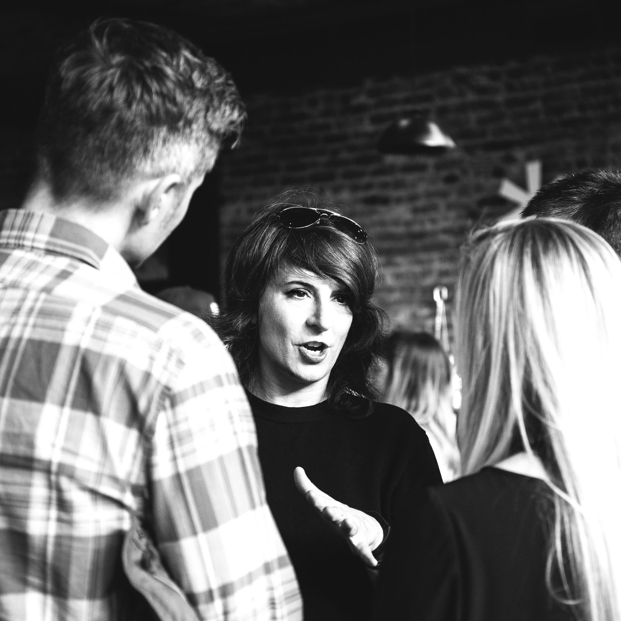 Black and white photo of a woman speaking, surrounded by people, with a man in a checkered shirt in the foreground and a person with striped clothing in the lower left corner.