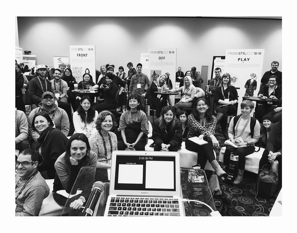 A group of diverse people at a conference or seminar, sitting and standing in front of posters, with a laptop and microphone in the foreground.