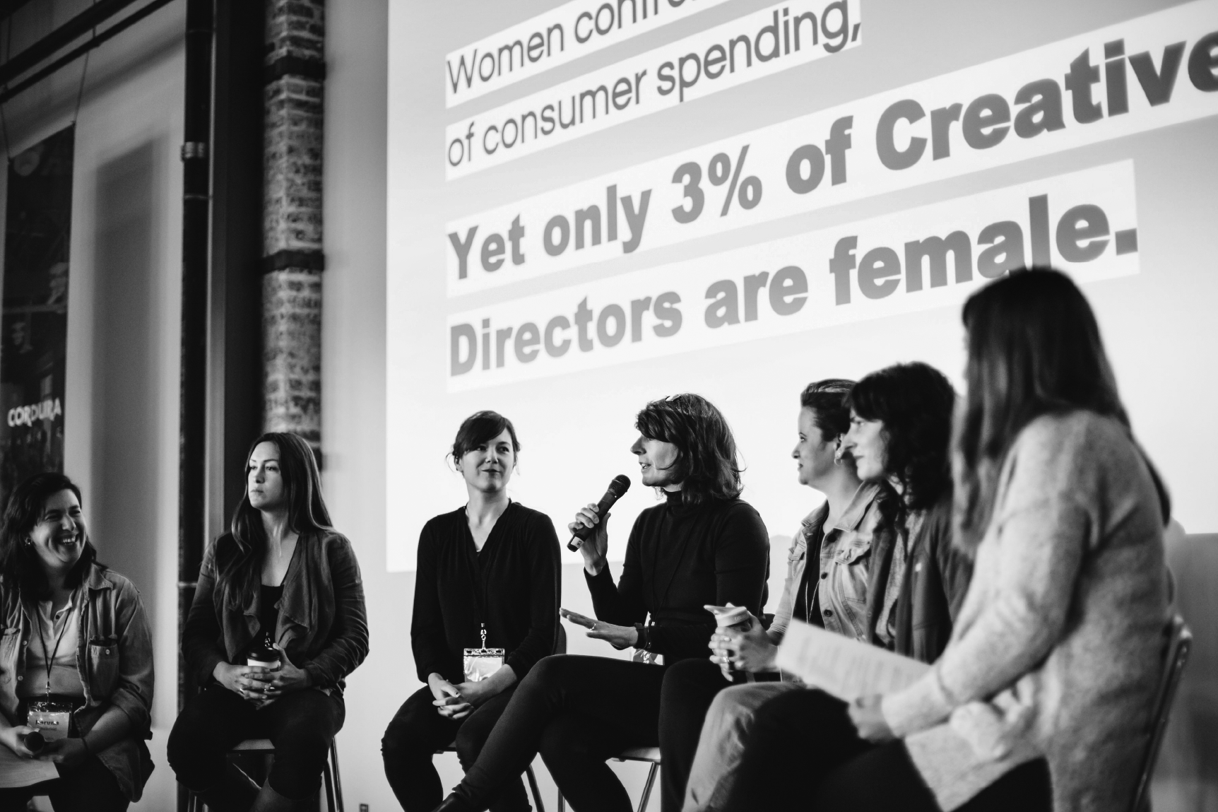 Group of women sitting on a stage participating in panel discussion or presentation at a conference, with a large screen behind them displaying text about women in creative director roles.