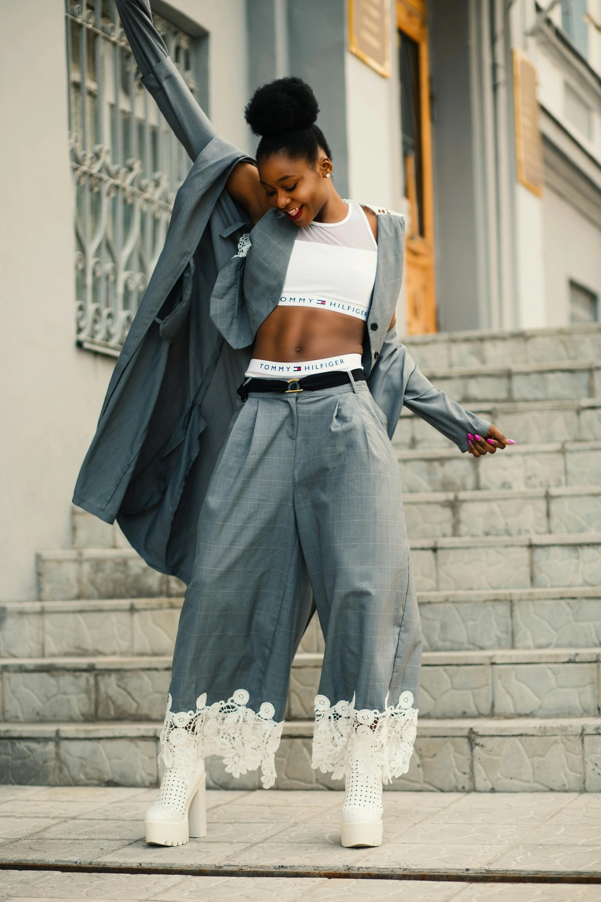Black woman with natural hair and a joyful expression dances on stone steps, wearing a stylish grey outfit with lace-trimmed pants and platform boots.