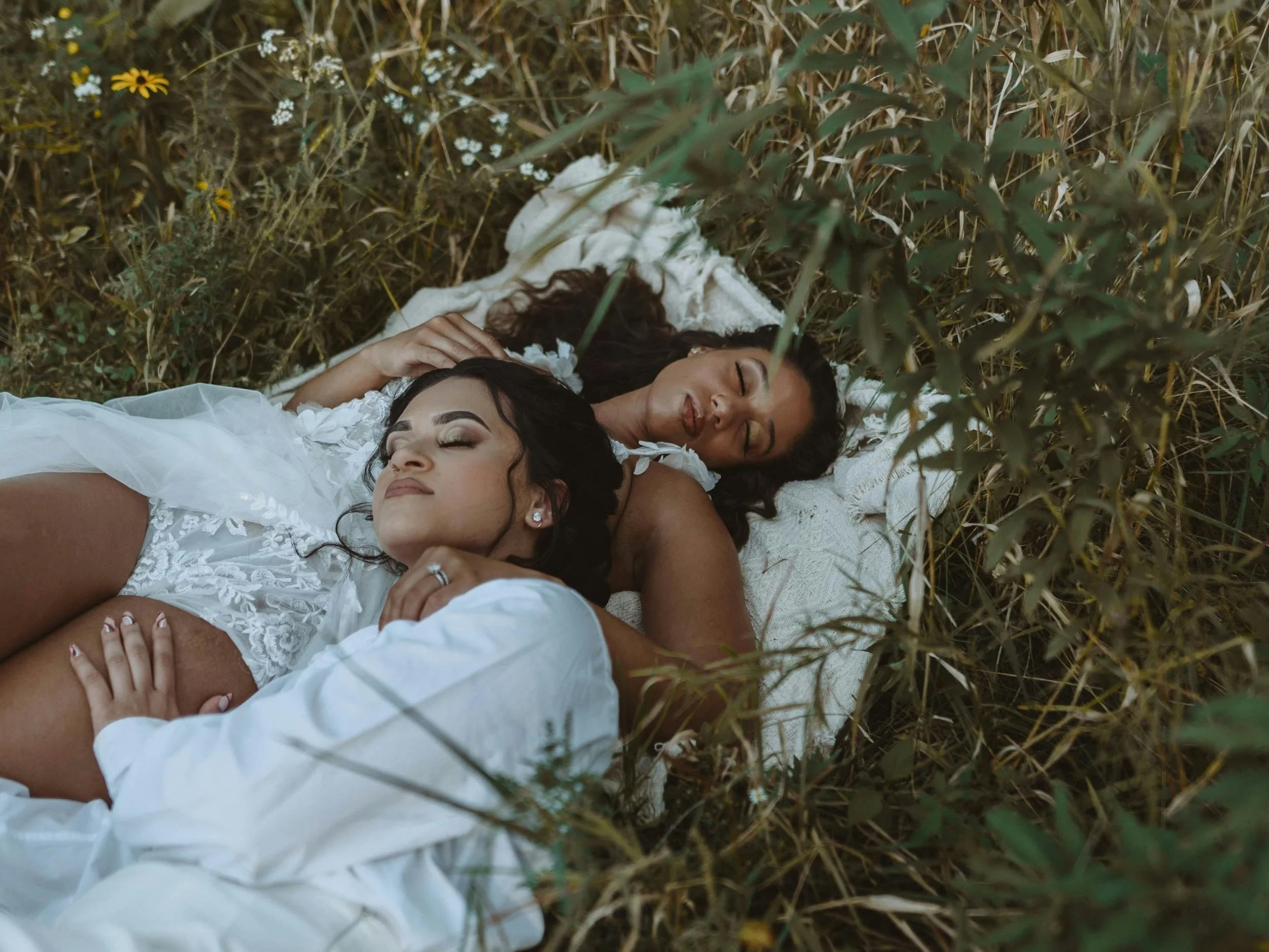Two women lay peacefully together on a blanket in a field of tall grass and wildflowers, eyes closed and bodies gently intertwined. One woman wears a white shirt while the other wears a lace dress, both surrounded by nature, intimacy, and calm.