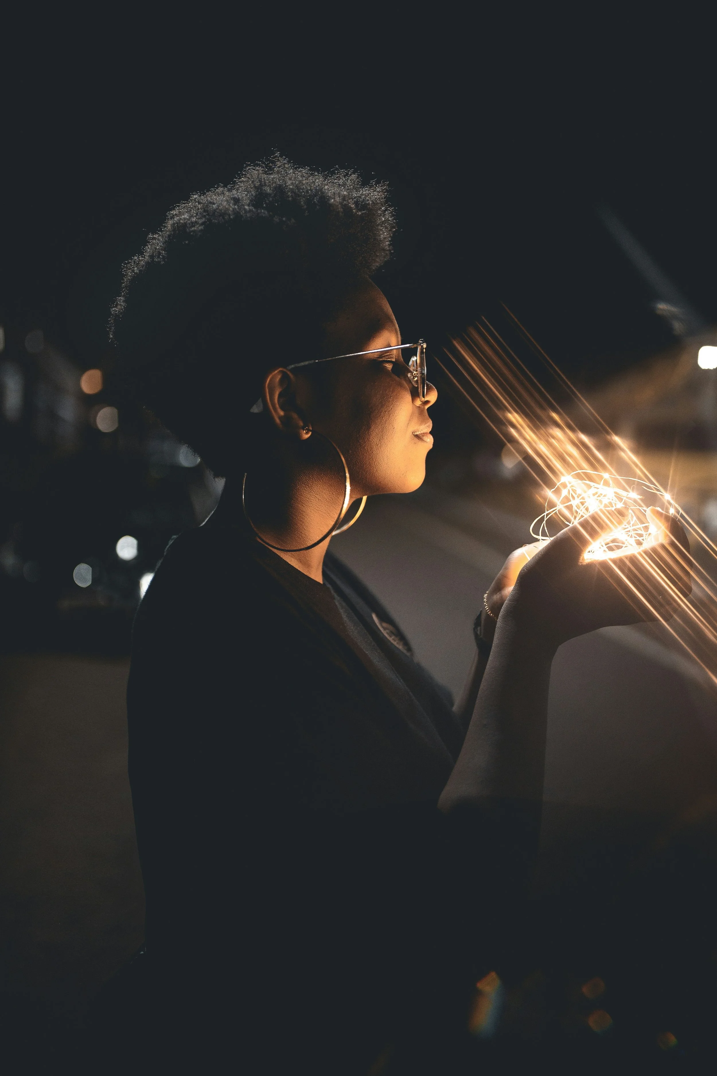 A Black woman with an afro hairstyle and glasses stands at night, holding glowing string lights in her hands. The lights cast a warm glow on her face, creating a serene and magical atmosphere.