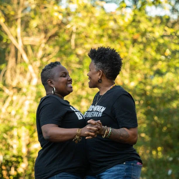 Two Black women stand outdoors, holding hands and smiling at each other. They are wearing matching black t-shirts and jeans, sharing a joyful and intimate moment in a natural setting with greenery in the background.