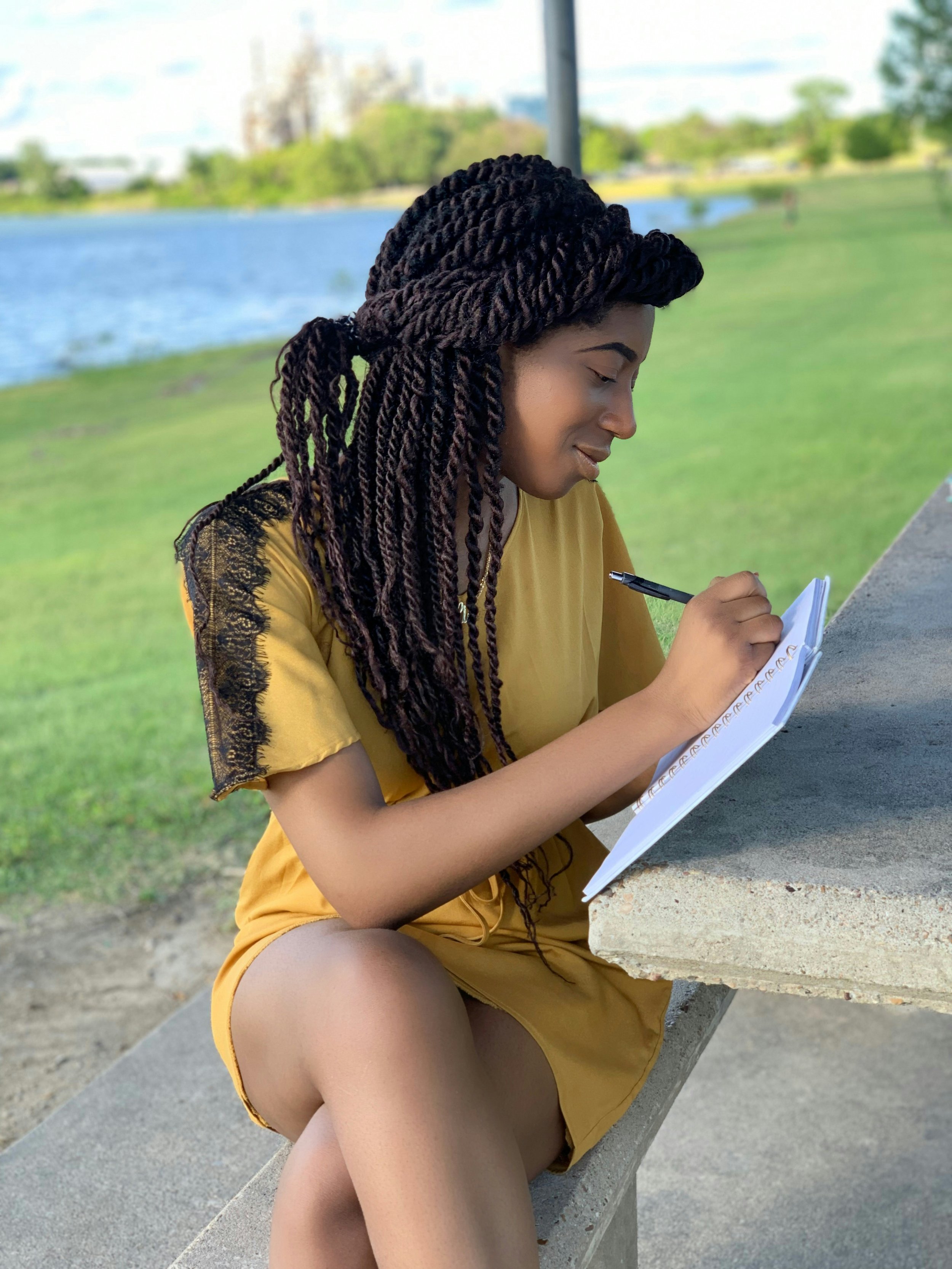Black woman with long twists journaling at a concrete picnic table by the water, wearing a mustard yellow dress, deeply focused and smiling softly as she writes.