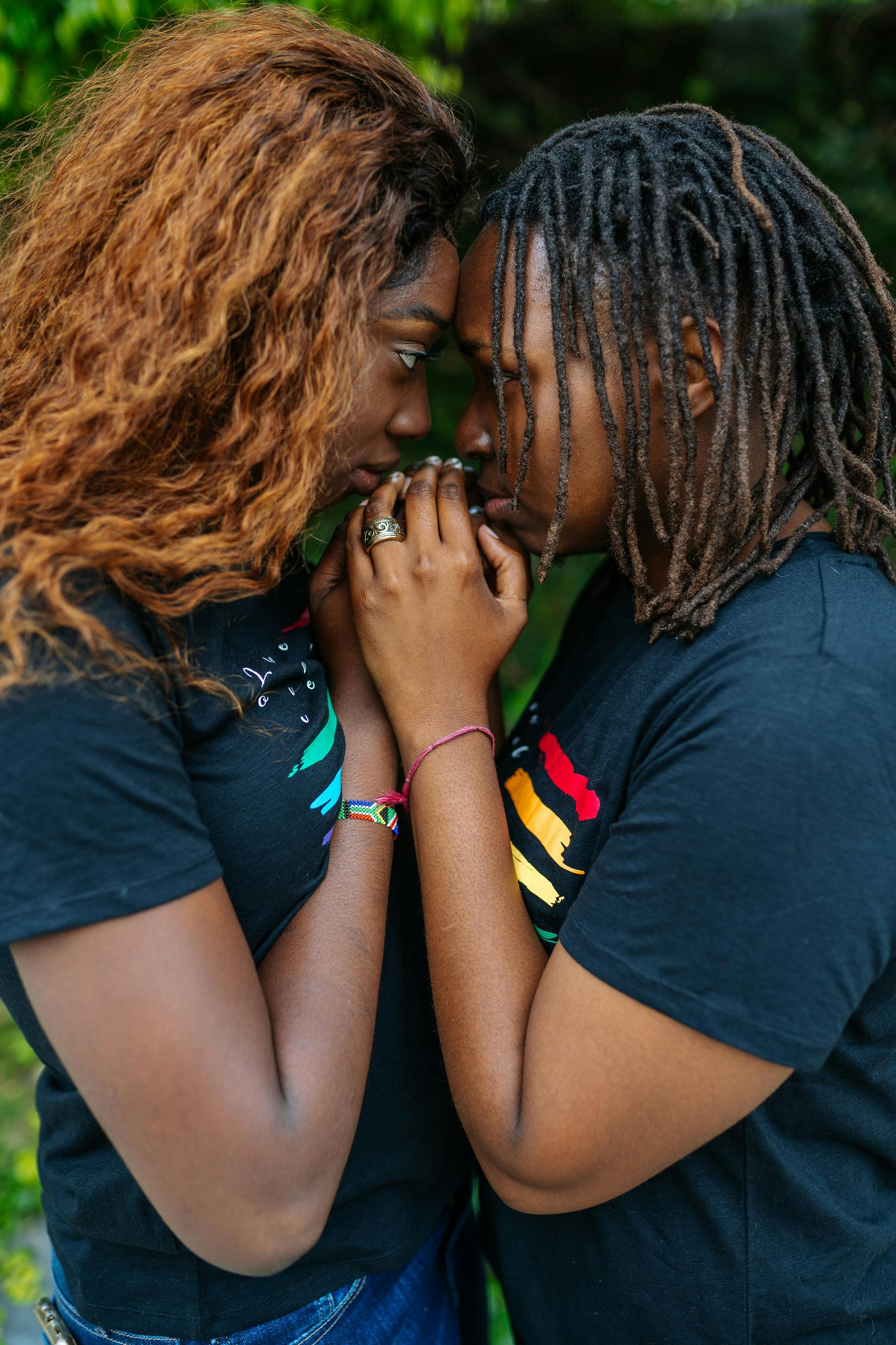 Two African American women standing close together, holding hands and touching their foreheads, sharing an intimate and supportive moment. Both women are wearing black t-shirts with colorful designs and are set against a background of lush greenery,