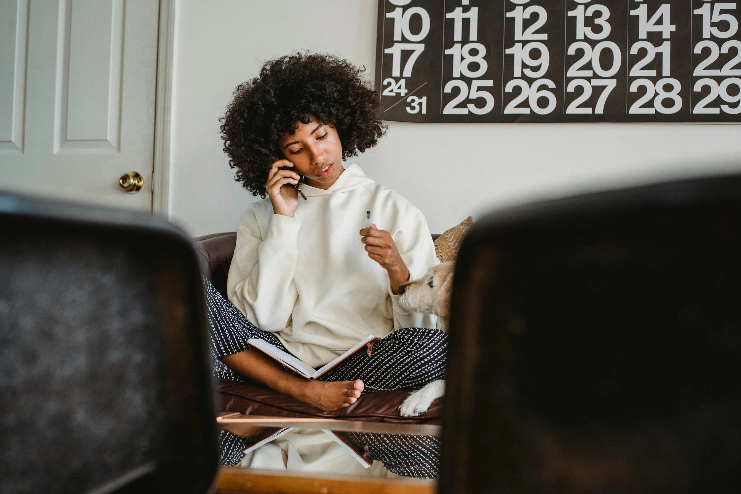 An African American woman sits comfortably on a couch, engaged in a phone conversation with a pen and notebook in hand. A calendar is visible on the wall in the background. This setting highlights the convenience and flexibility of online counseling