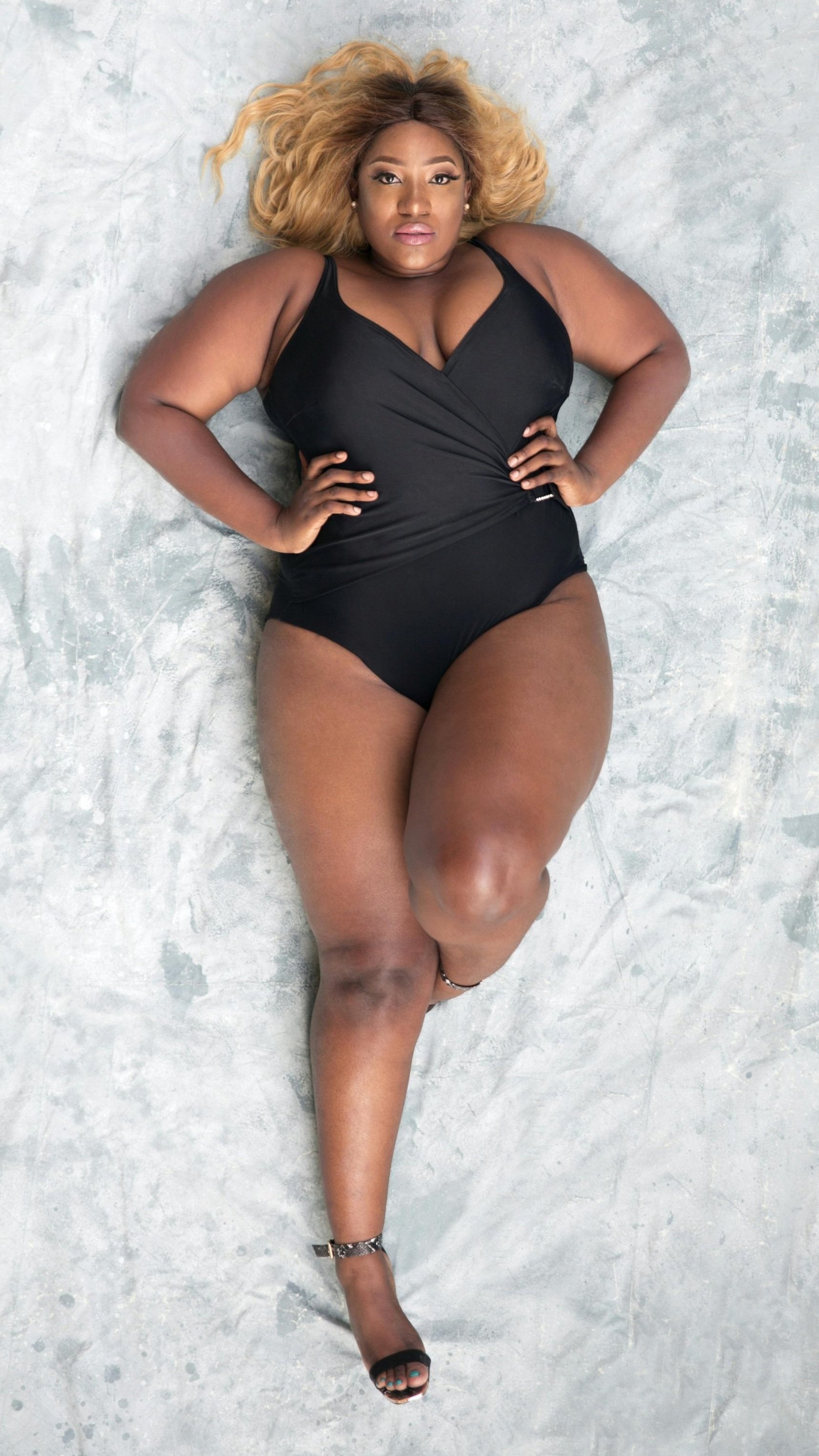 A black woman lying confidently on a bed in a black bodysuit, radiating strength and softness. Her body language exudes an unapologetic presence, reflecting empowerment and self-acceptance.