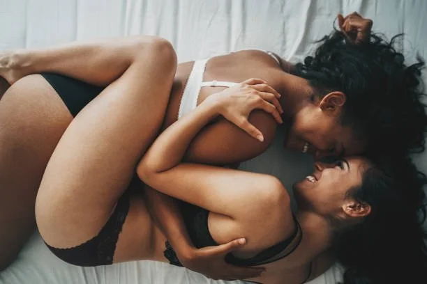 Two women of color lie intertwined on a bed, laughing and holding each other with joy and intimacy, capturing a moment of playful connection and deep affection.