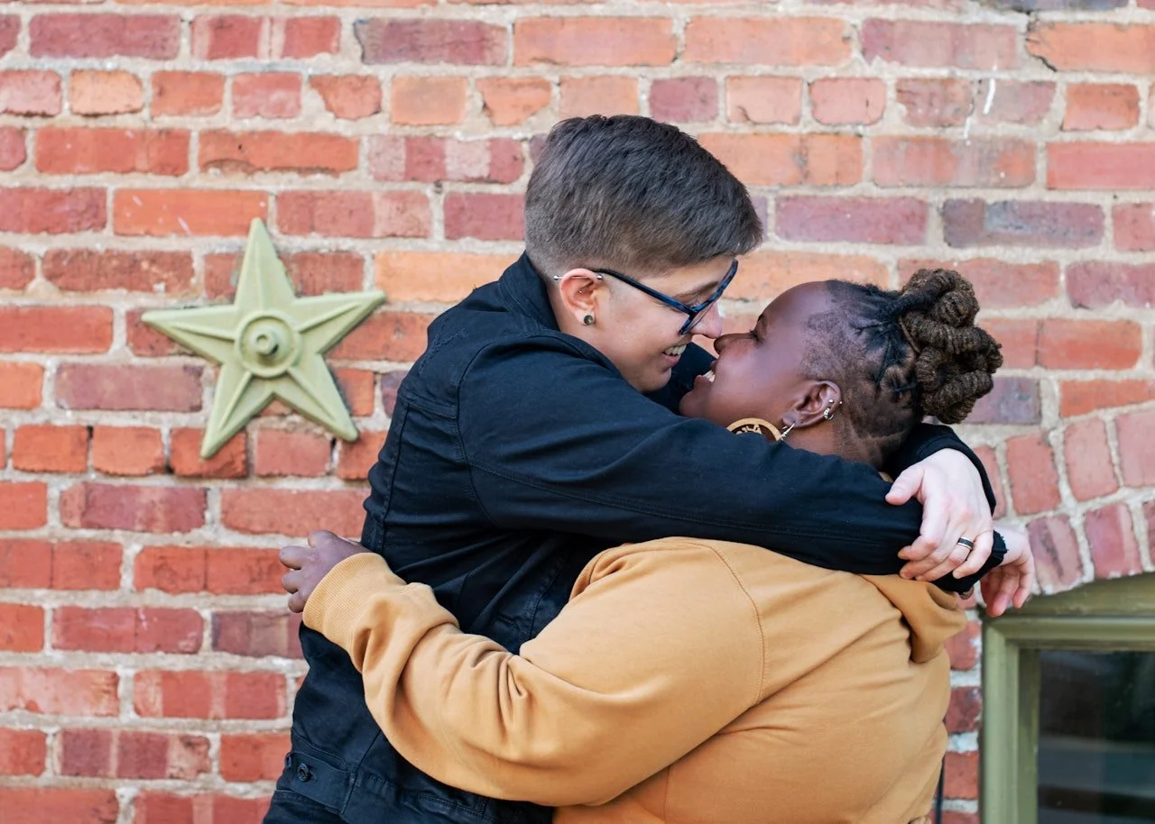 Queer interracial couple embracing with joy in front of a brick wall, expressing intimacy, safety, and deep connection.