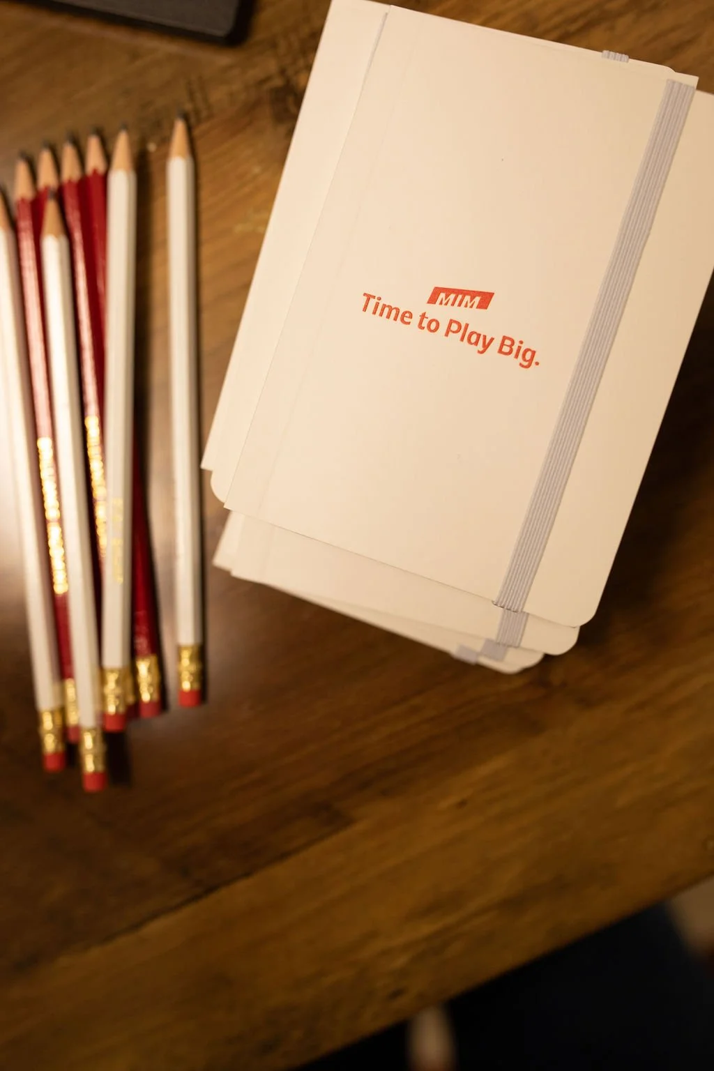 Stack of white notebooks or notepads with red printed text that reads 'MIM Time to Play Big.' on top of a wooden surface with a row of red, white, and gold pens to the left.