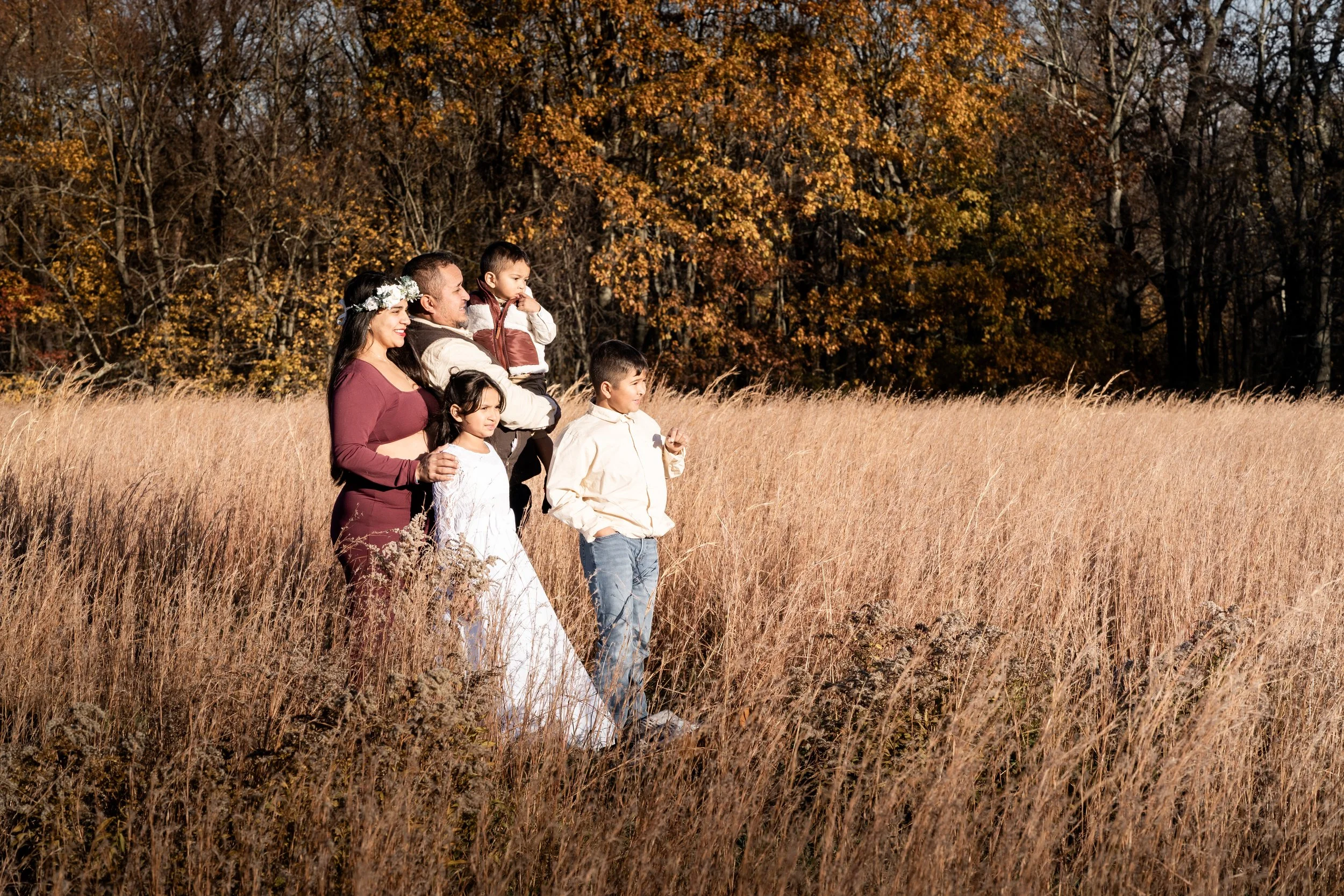 A family of six standing in a field of tall, dry grass during autumn, with a background of trees with orange and yellow leaves.