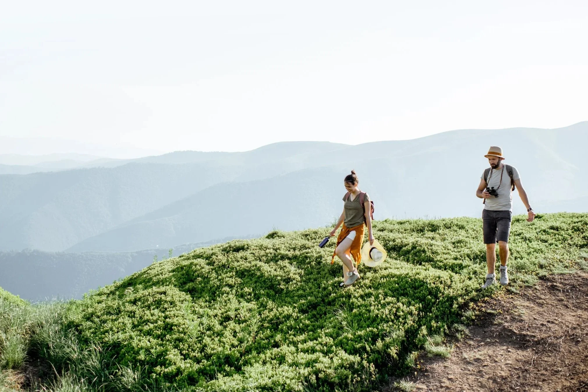 A man and woman hiking through a grassy mountain trail on a bright, clear day with mountains in the background.