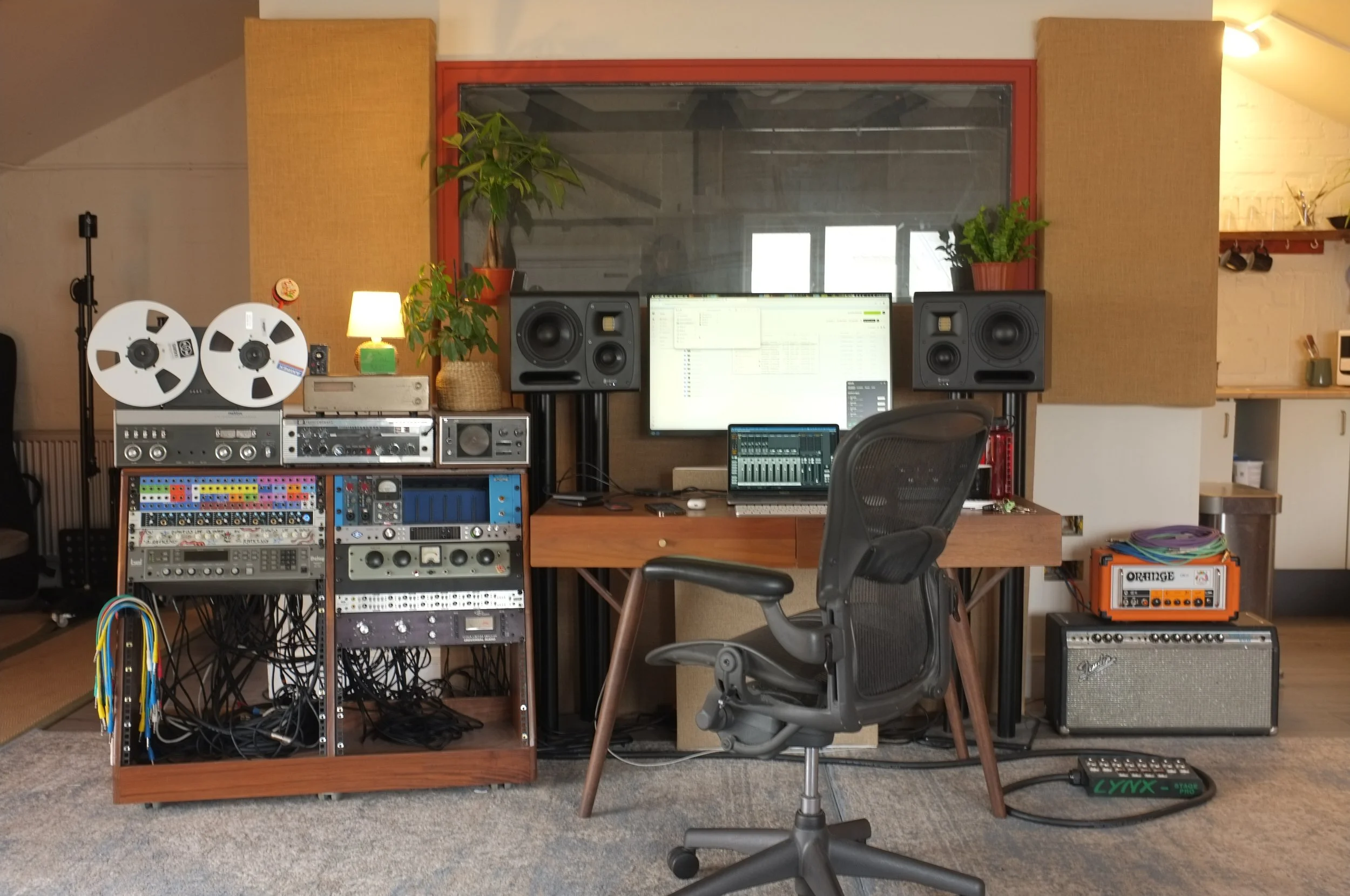 A music studio workspace with various audio equipment, a computer monitor, studio speakers, a chair, and plants on the window sill.