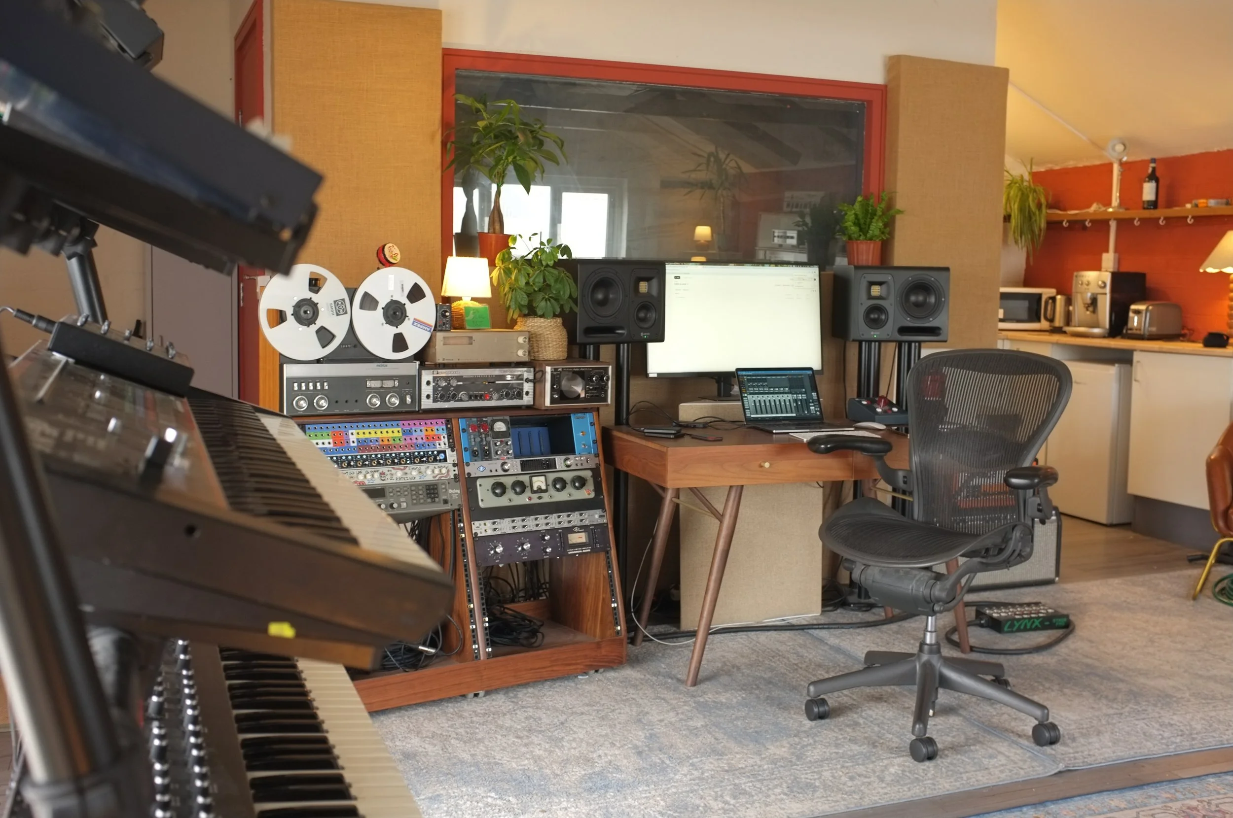Music studio with a keyboard in the foreground, mixing console, large speakers, a computer monitor, a small tablet, potted plants, and various audio equipment on shelves. A cushioned chair is in front of the setup, with a gray carpet on the floor.