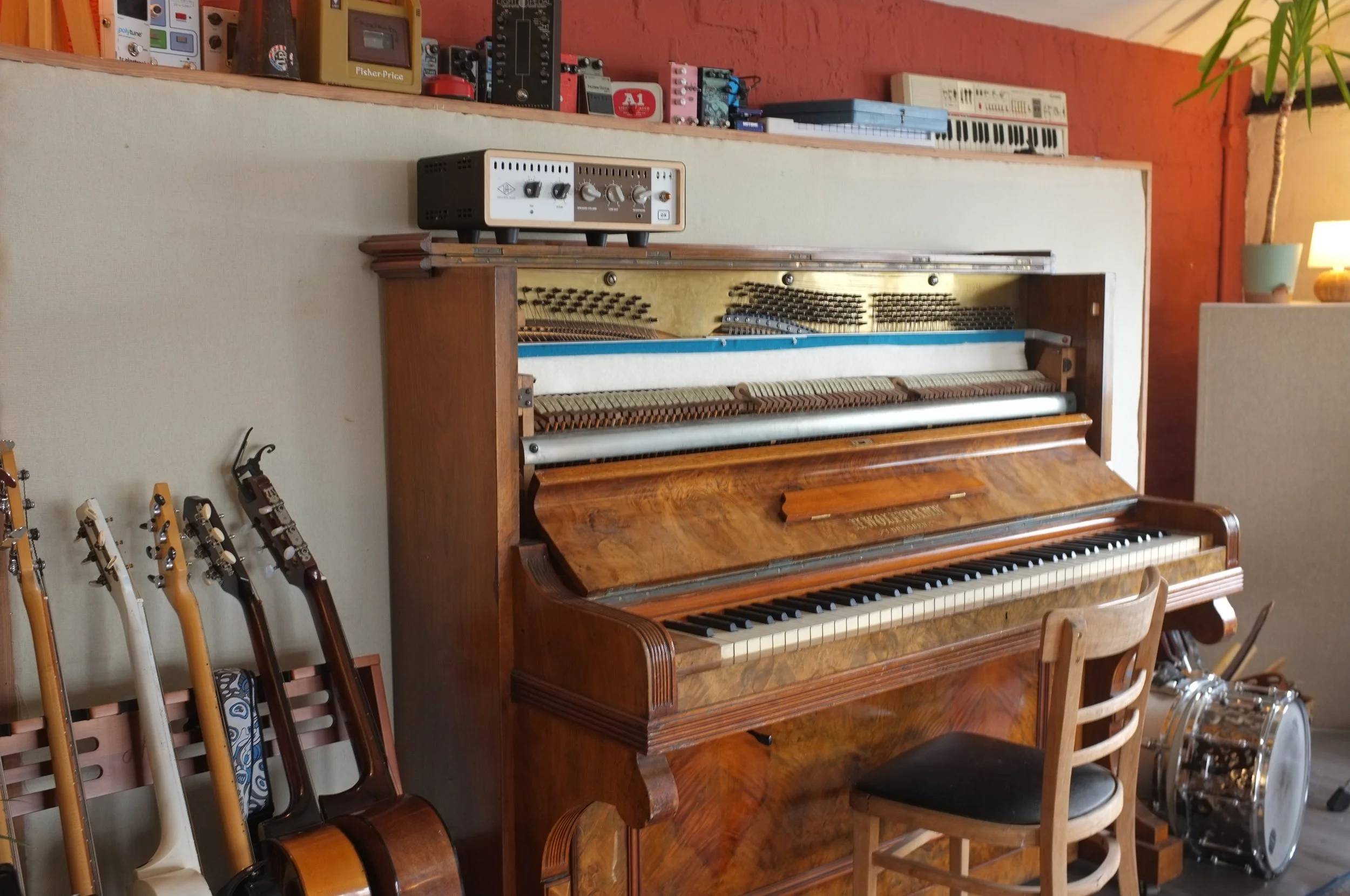 A wooden upright piano with its keys exposed, located in a cozy room. To the left of the piano, multiple guitar necks are stacked on a stand. On top of the piano, there is a small vintage stereo or amplifier. Behind the piano, a shelf holds an electronic keyboard, various effects pedals, and other musical equipment. To the right, a lamp with a green shade and a tall plant are visible.