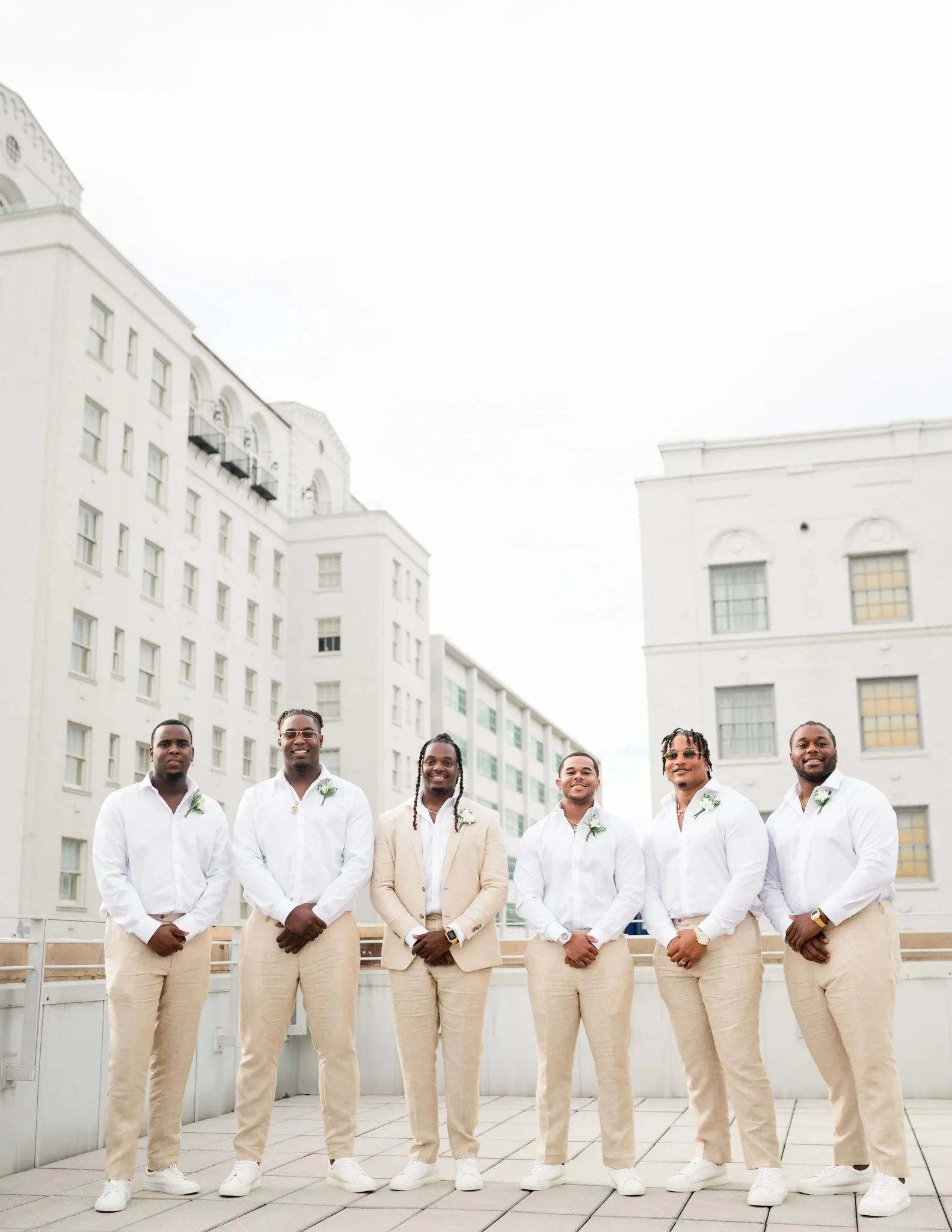 Six men dressed in white shirts and beige pants standing on a rooftop in front of white buildings, smiling at the camera.