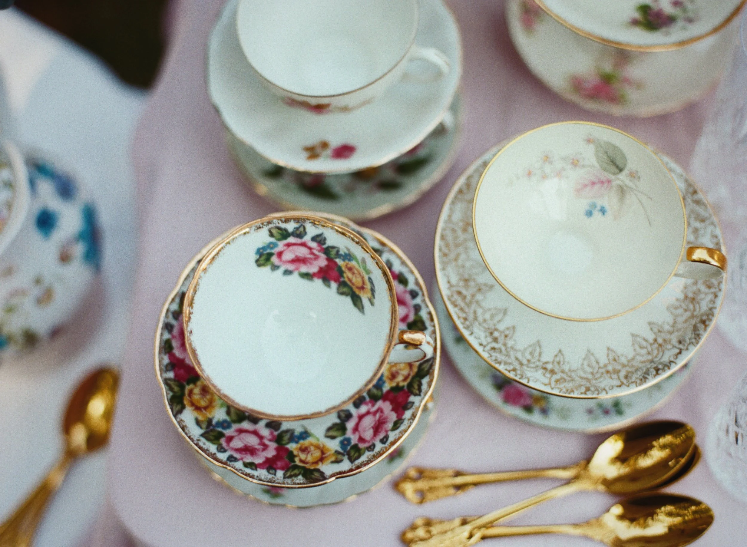 A collection of fine china teacups and saucers with floral and gold patterns, placed on a pink tablecloth, alongside gold spoons.