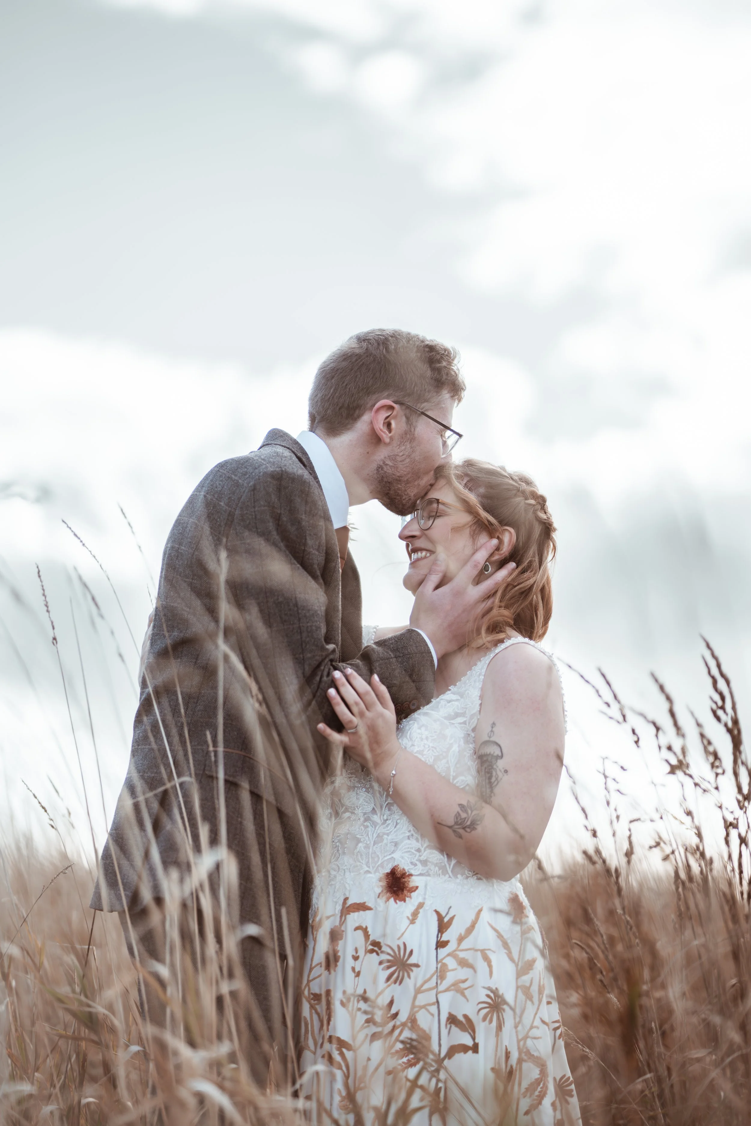 A couple embracing in a field of tall grass, with the man kissing the woman's forehead, both smiling and wearing wedding attire.