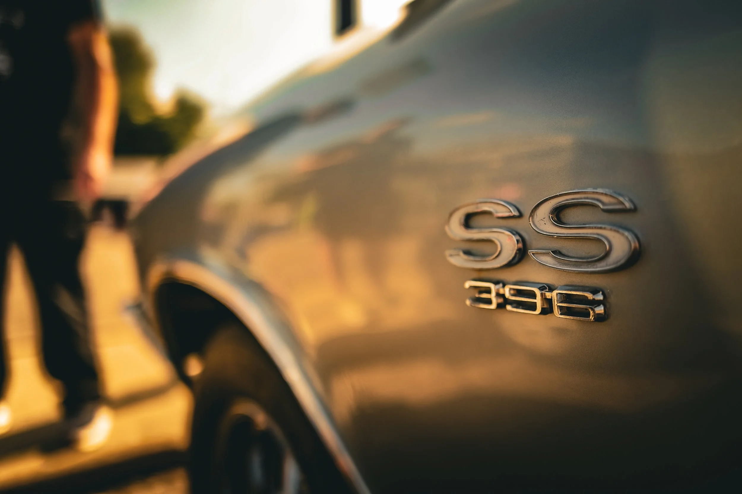 Close-up of a silver car badge with the number 396 on a black car body, reflected in the sunset light.