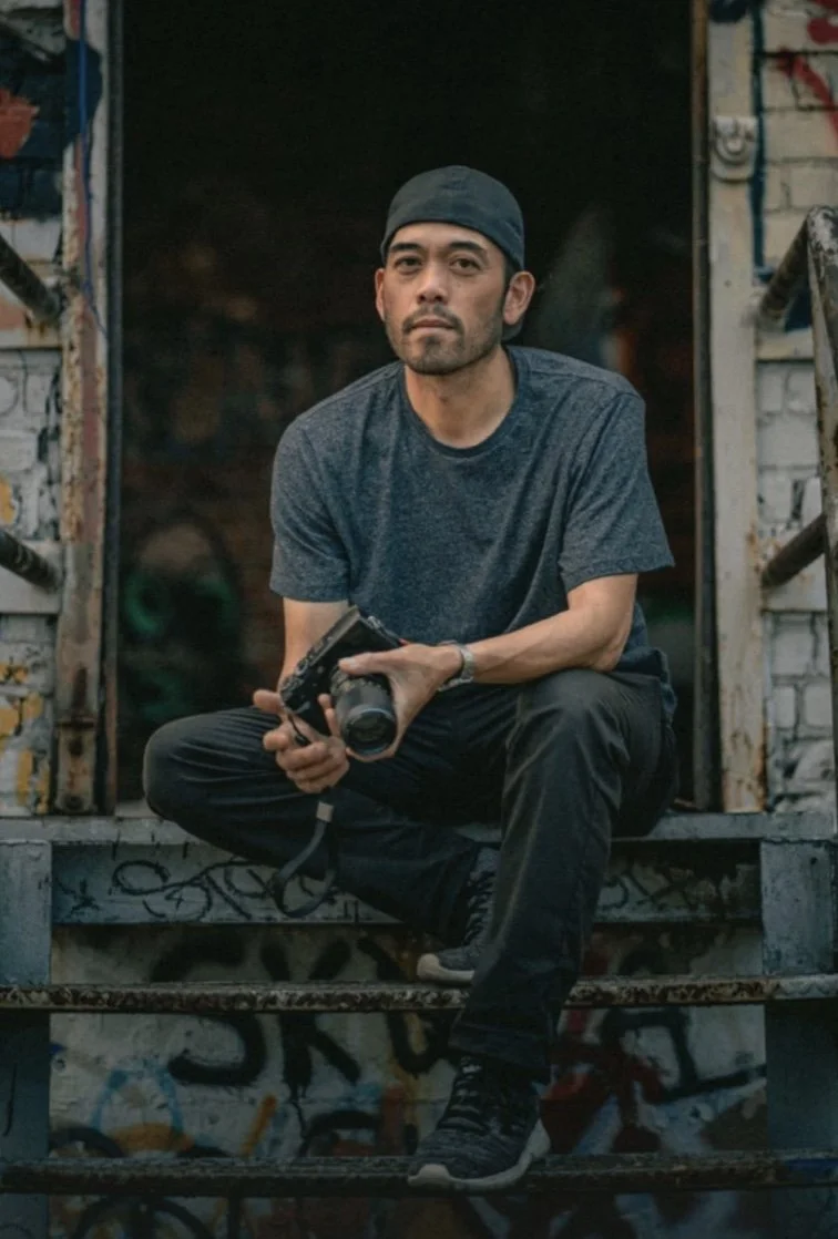 A man sitting on graffiti-covered metal stairs in front of a dark background, holding a camera, wearing a gray t-shirt, black pants, black shoes, and a cap.