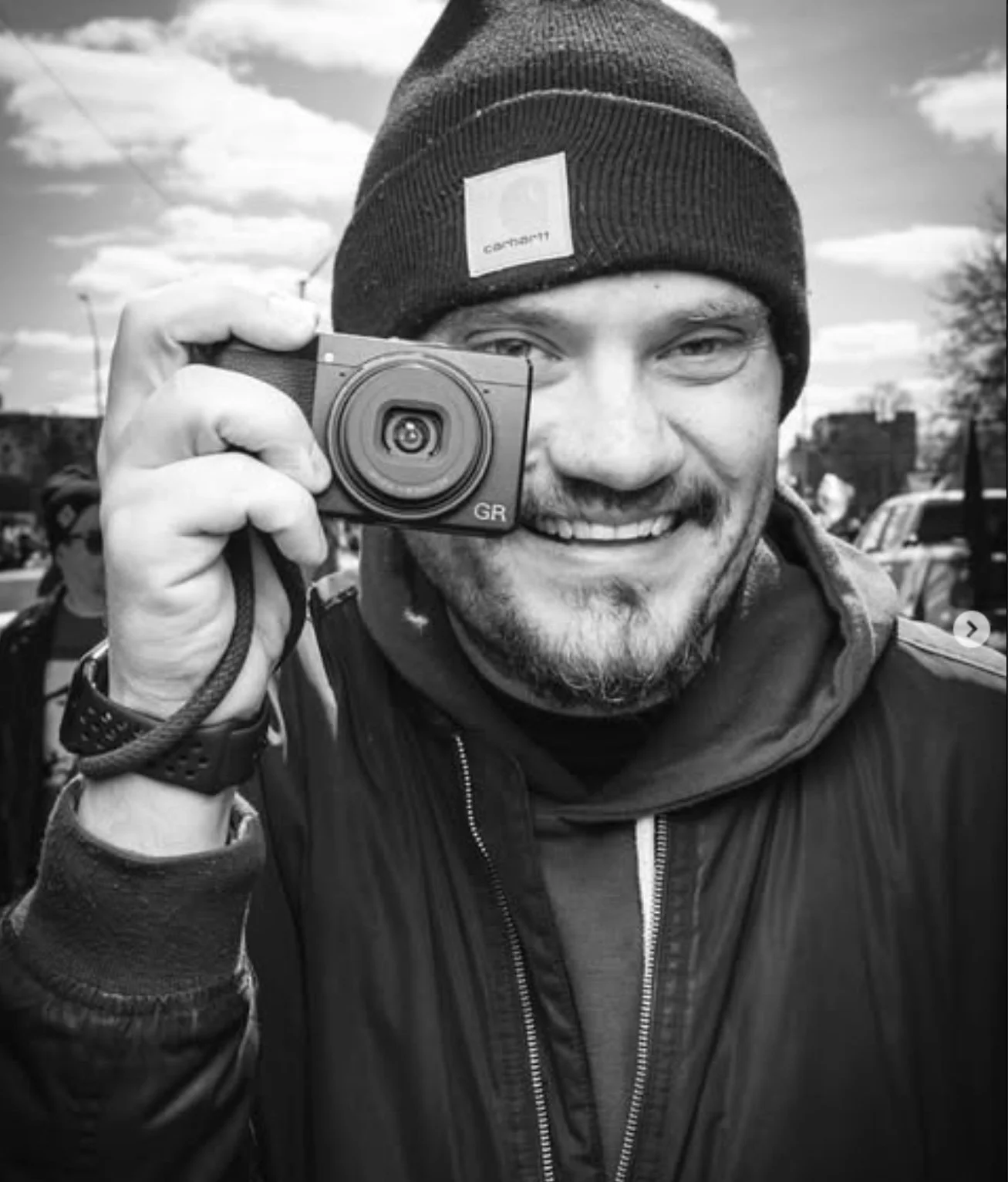 A smiling man wearing a black beanie and a dark jacket, holding up a small digital camera to his face outdoors, with a cloudy sky and some blurred people and cars in the background.