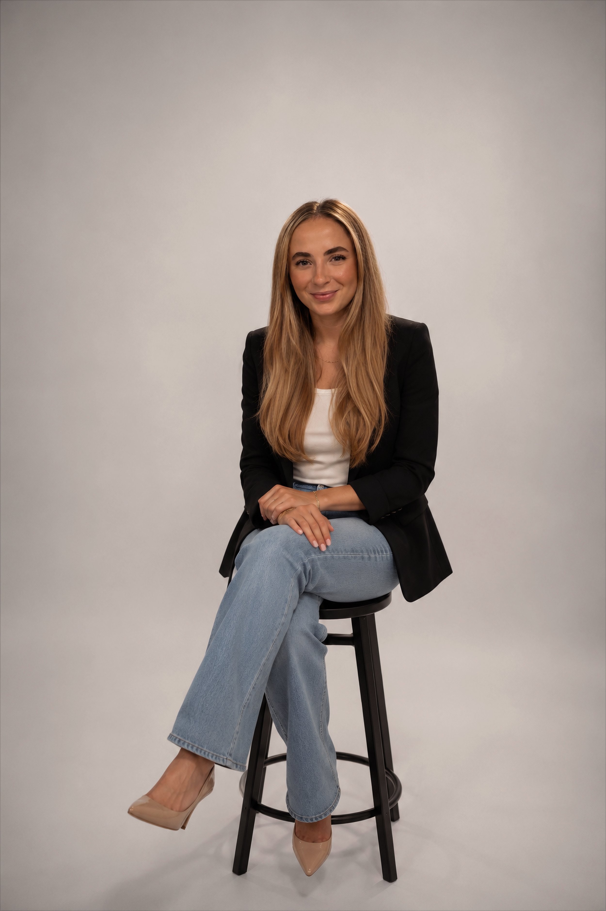 A young woman with long blonde hair sitting on a black bar stool against a plain gray background. She is wearing a black blazer, white top, light blue jeans, and beige high heels. She has a slight smile and is looking directly at the camera.